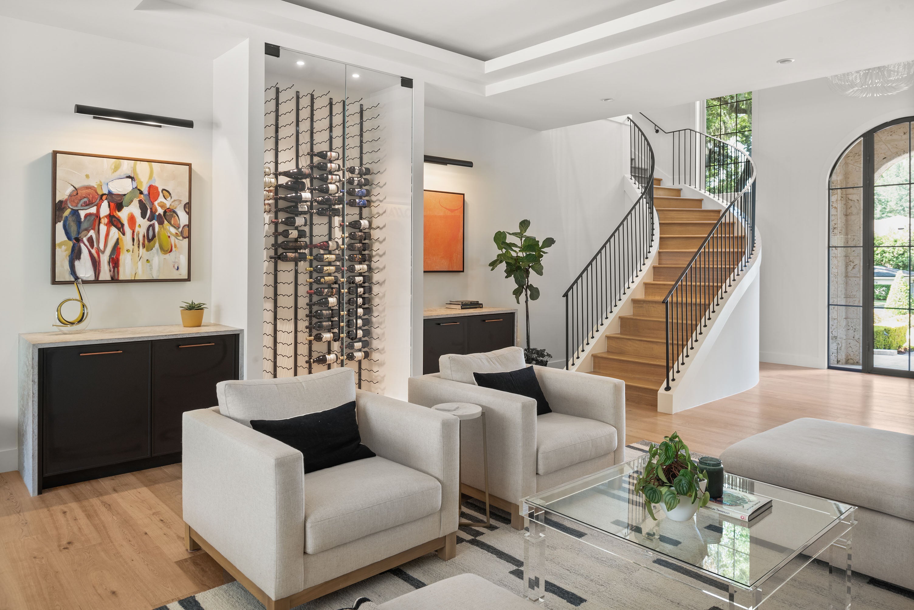 Modern living room with white chairs, a glass coffee table, and a staircase in the background featuring a wine cellar behind glass